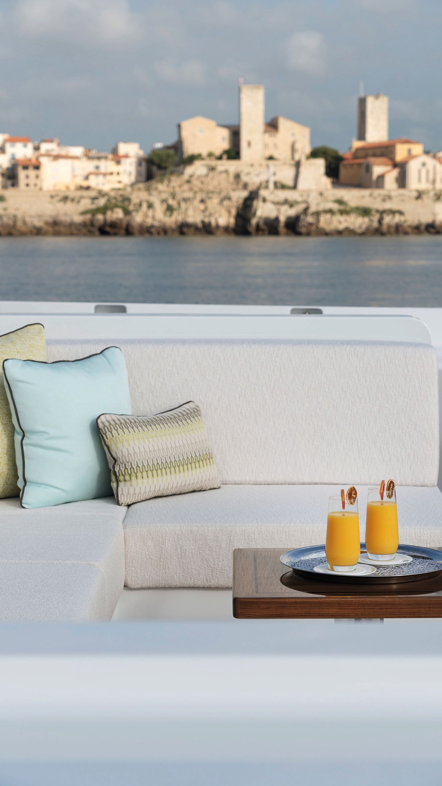 The corner of a sofa on an outdoor deck. In the background is a city skyline and in the foreground are two glasses of orange juice on a silver tray