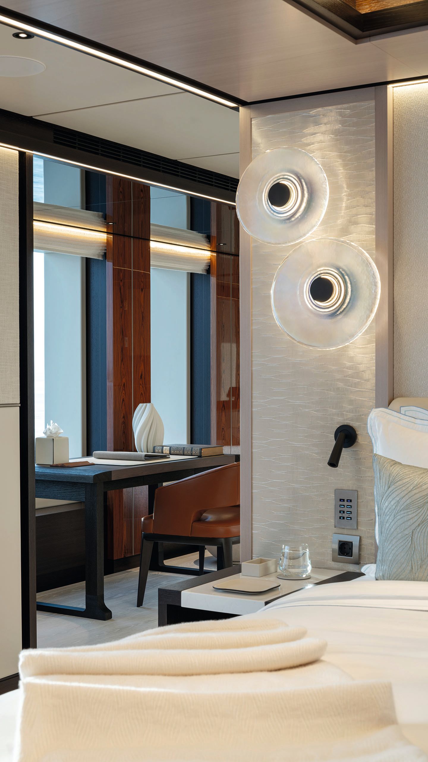 Partial view of the side of a bed looking towards a desk by a window