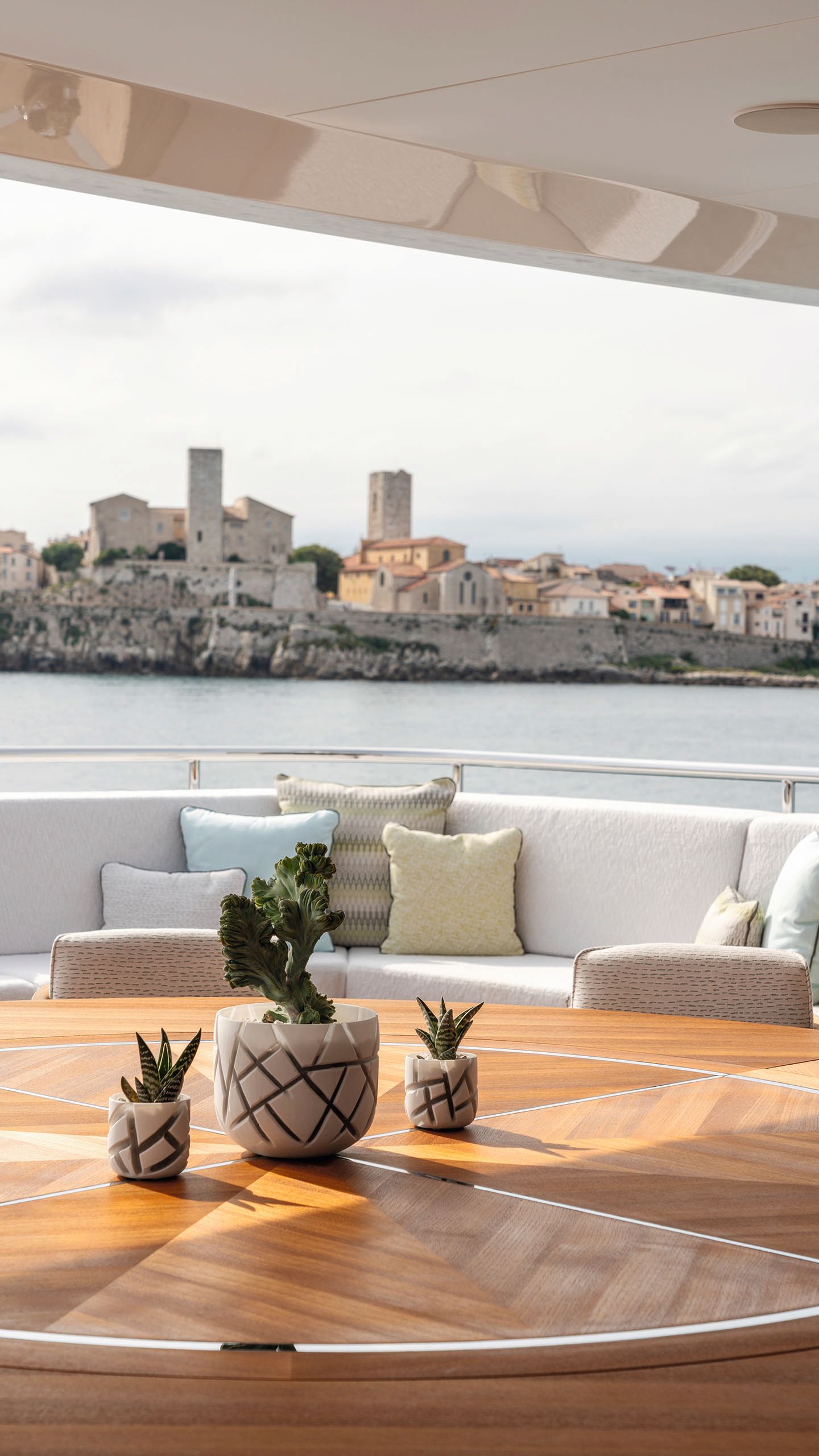 A round table with patterned top on a partially covered outdoor deck. There are three cacti in pots in the centre and a curved sofa and skyline in the background