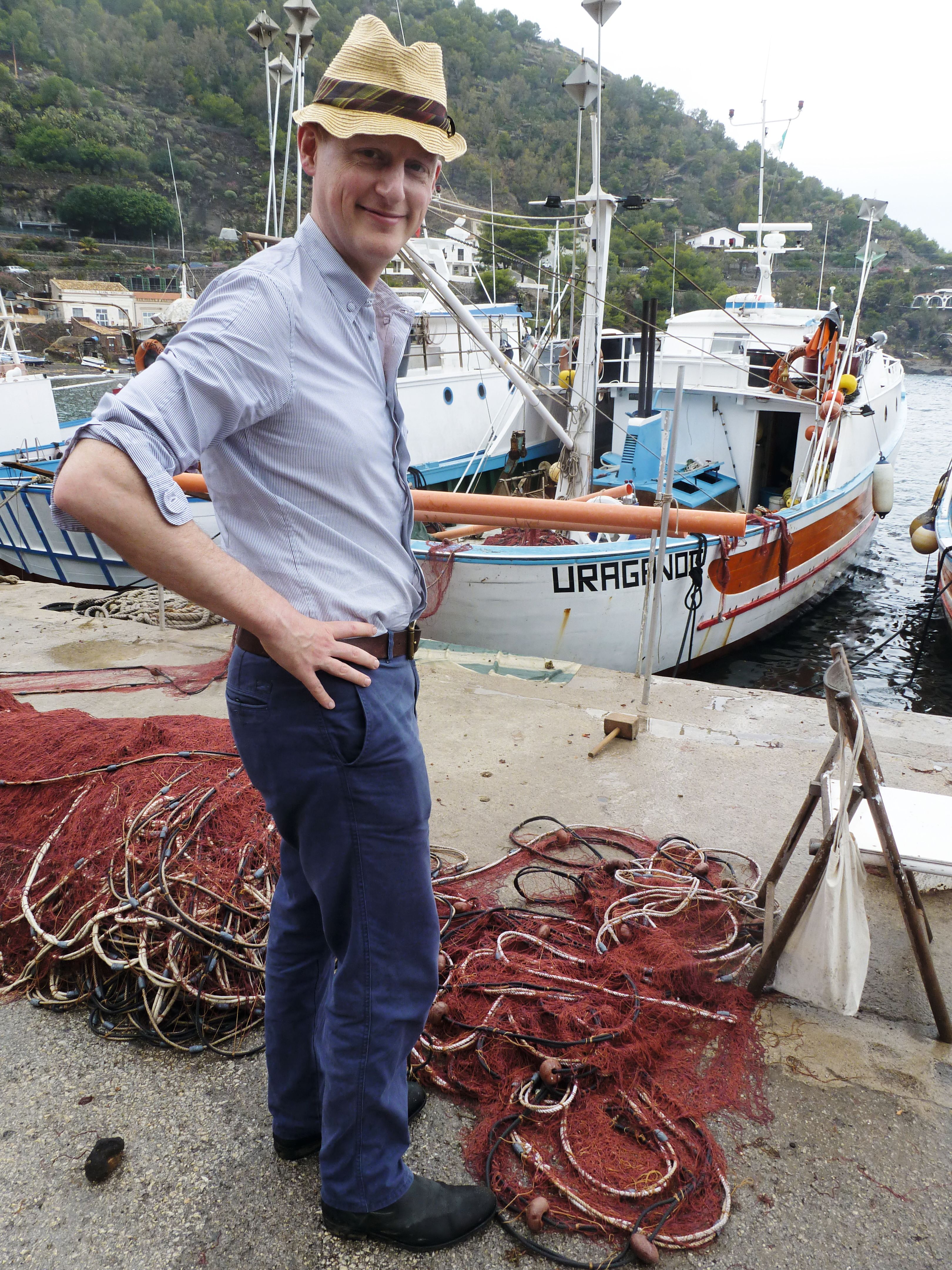 Harry Mount is standing on the side of a harbour with his hands on his hips. He's wearing a pale blue shirt with the sleeves rolled up and navy trousers. He has a straw hat on his head.