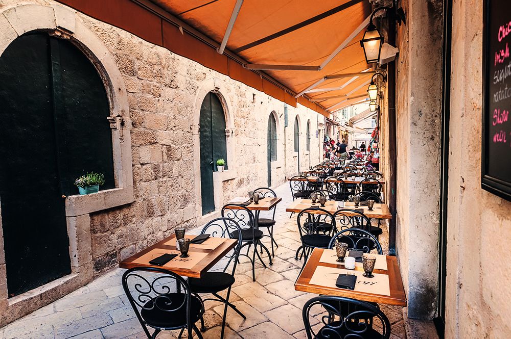 A courtyard with wrought iron chairs and tables outside a restaurant. There is an orange canopy above and the buildings are made from cream stone