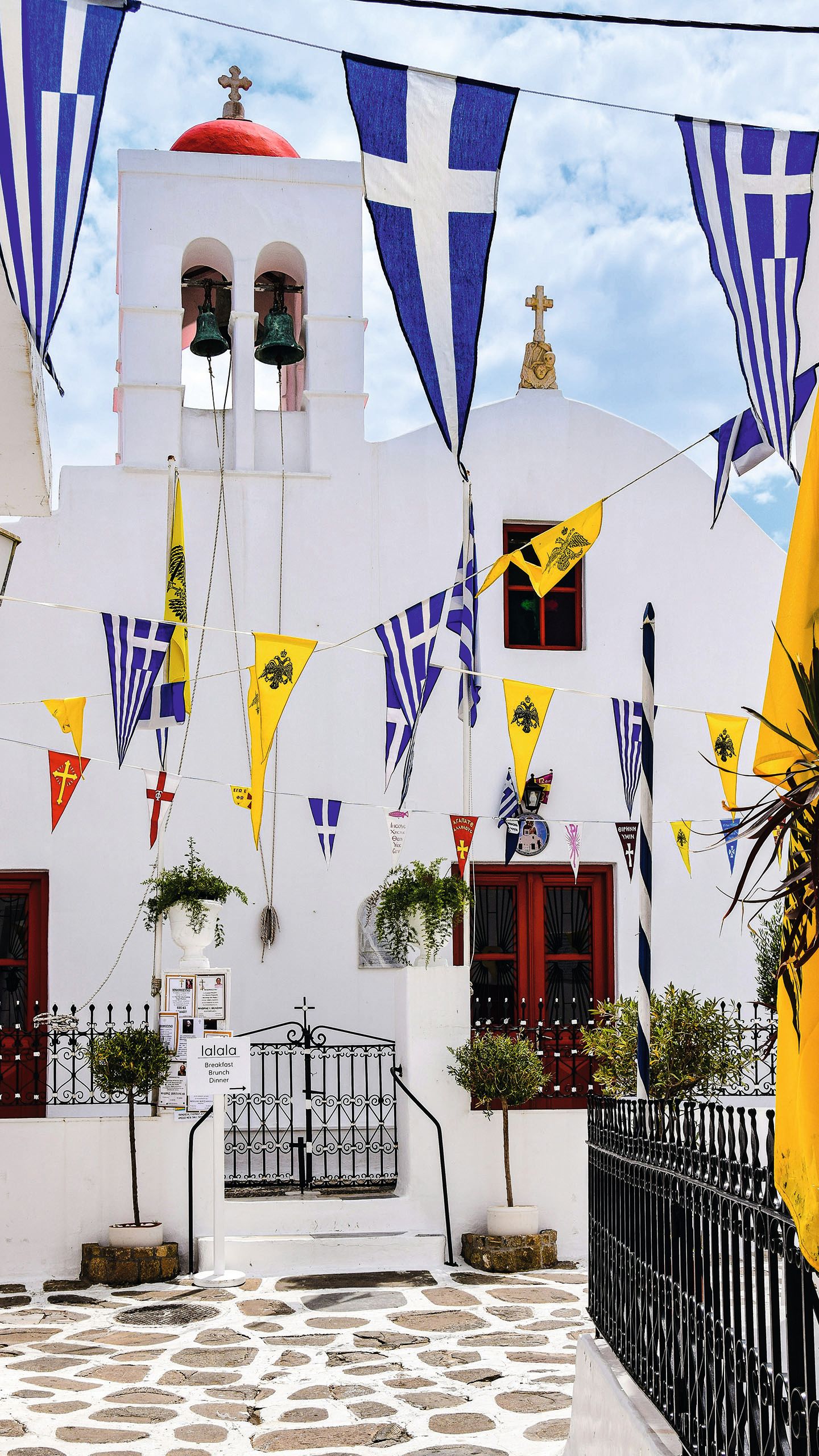 A white church in Mykonos with a red dome. There are strings of criss-crossing Greek flags leading up to it
