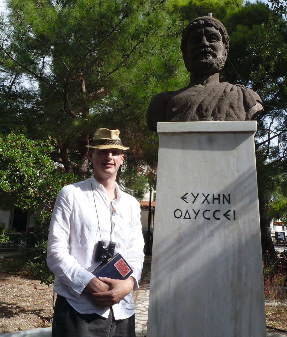 Harry Mount is wearing a long-sleeved, light white shirt and a straw hat. He is holding a navy blue book and has binoculars around his neck. The busy of Odysseus next to him has Greek writing on it