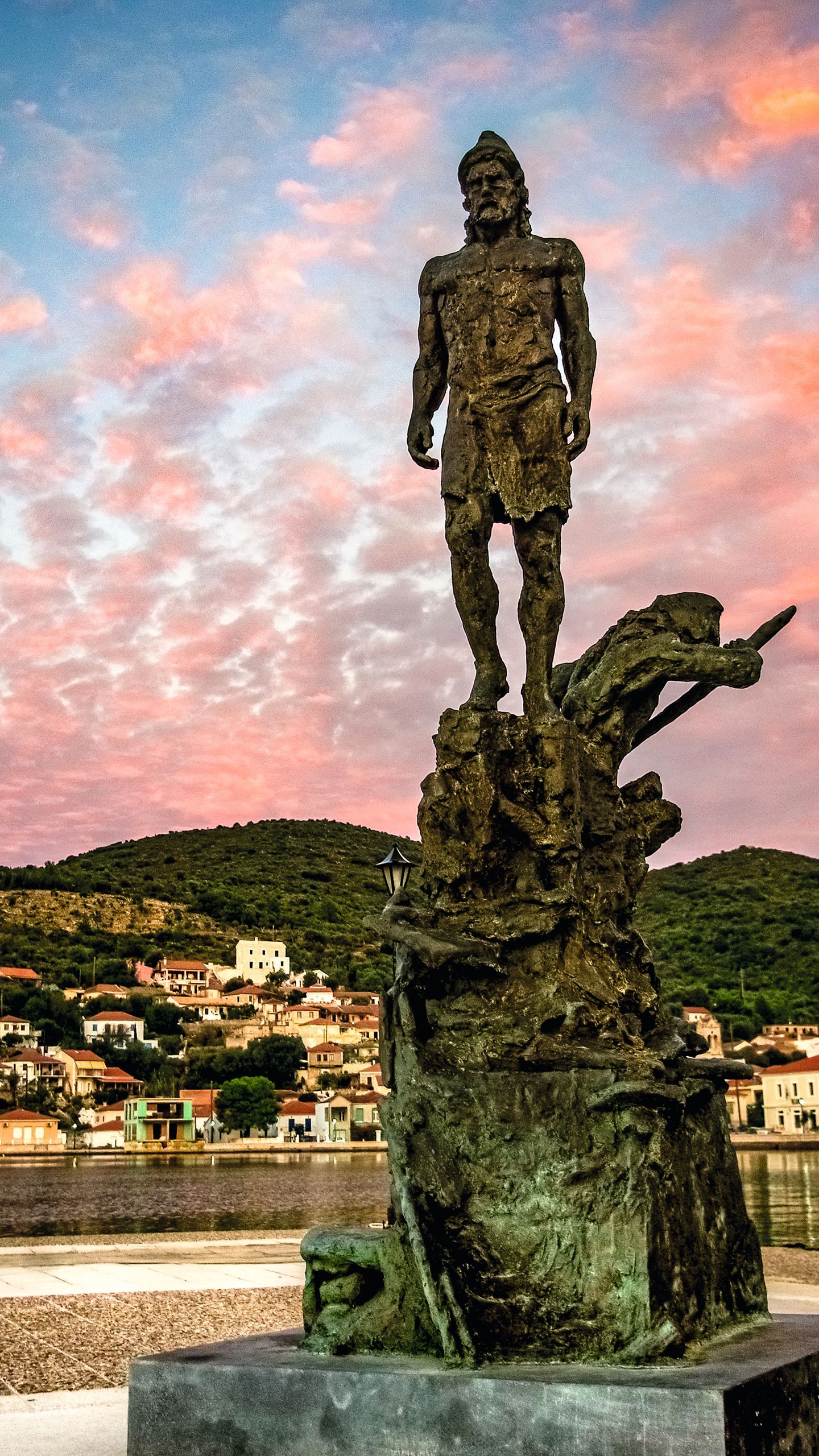 A statue of Odysseus - it is on a large stone plinth. Ithaca is behind and the sky is blue with pink clouds