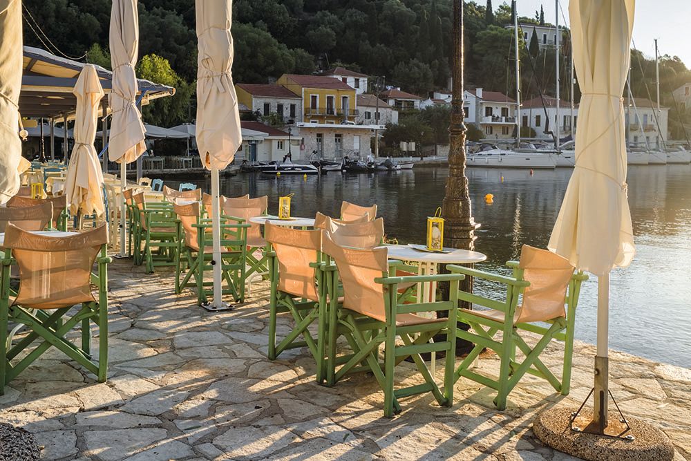 Tables and chairs on a dock at sunrise/sunset. The chairs are green wood with orangey seat covers and there are white parasols by the tables