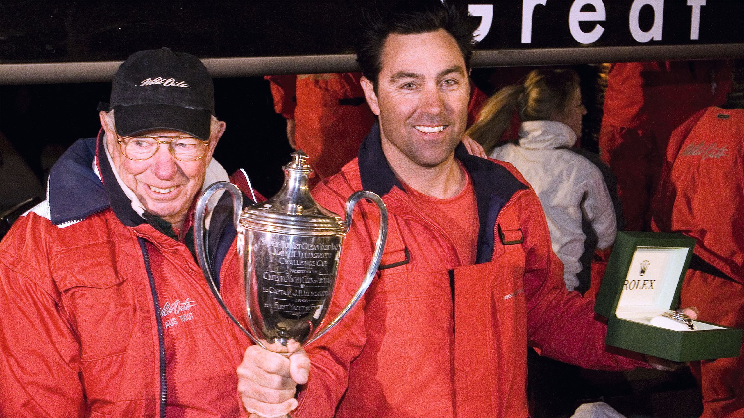 An older man standing next to a younger man holding a large silver trophy. They're both in red jackets