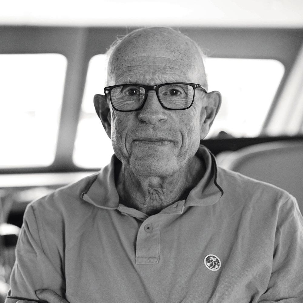 Black and white photo of Tom Whidden. He's looking at the camera but not smiling. He is wearing black-framed glasses, a polo short and is bald.
