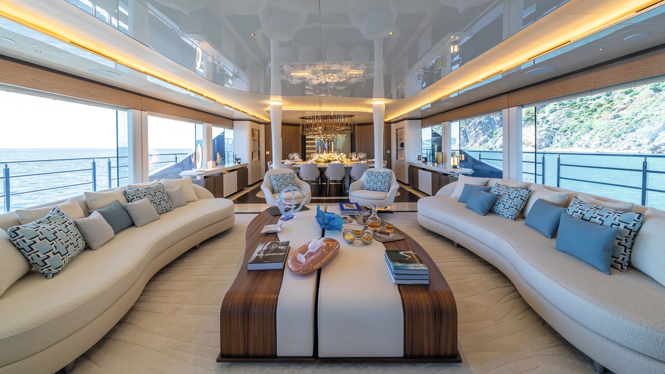 Wide view of the salon which features two long slightly curved sofas in neutral colours facing each other. A curved long coffee table sits in between with two armchairs at the end. At the back is the dining table with a chandelier above