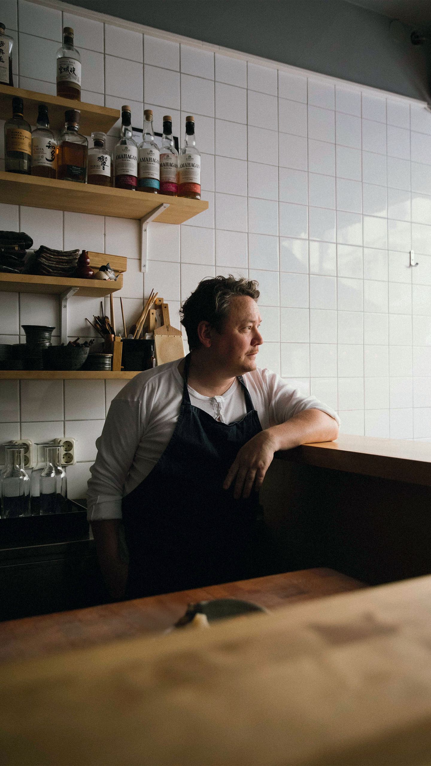 A man in a white t-shirt and navy blue apron. He has short blond hair and is standing behind a bar with his arm propped up on the counter. Behind him the wall is tiled white and there are shelves holding spirits, crockery and glassware