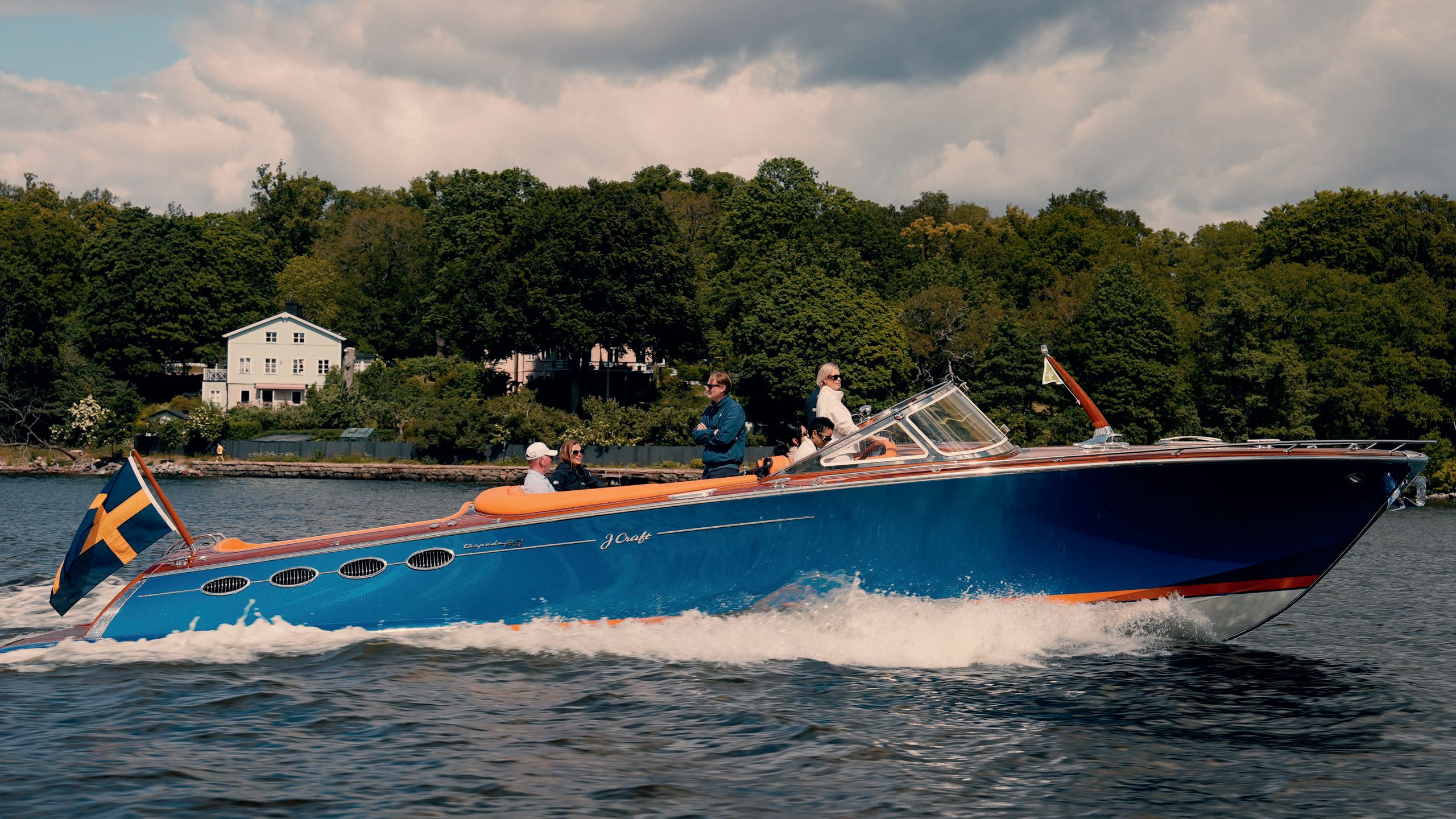 Side view of a J Craft vessel on the water. It's painted in blue, has an orange trim and a Swedish flag at the back