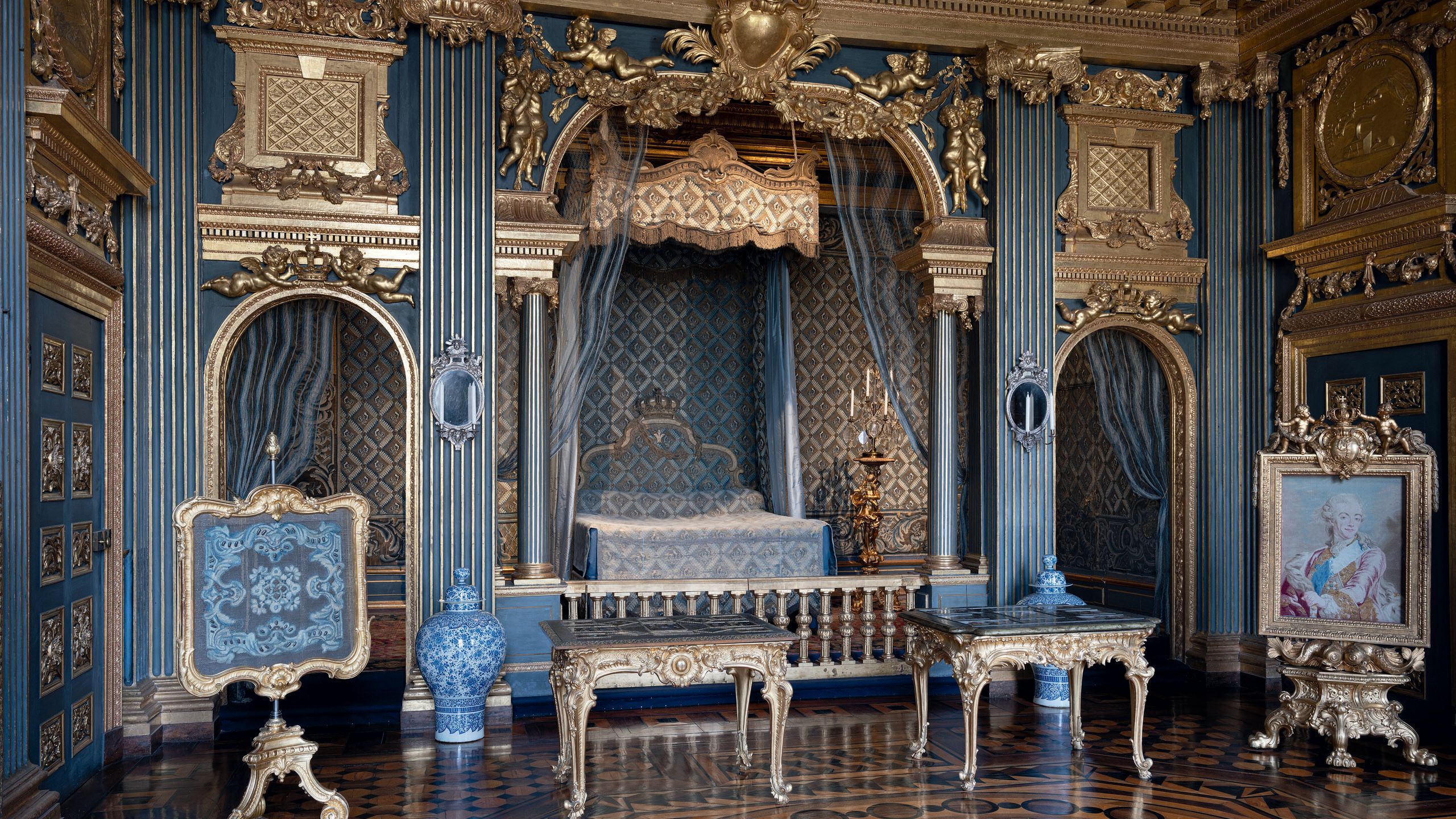 A bedroom with ornate decor in blues and golds. There is a bed in the background surrounded by drapes, blue and white vases on the floor and tables with filligree detail at the front