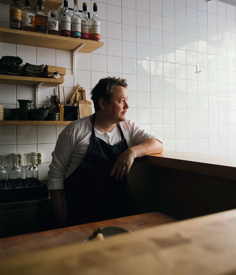A man in a white t-shirt and navy blue apron. He has short blond hair and is standing behind a bar with his arm propped up on the counter. Behind him the wall is tiled white and there are shelves holding spirits, crockery and glassware