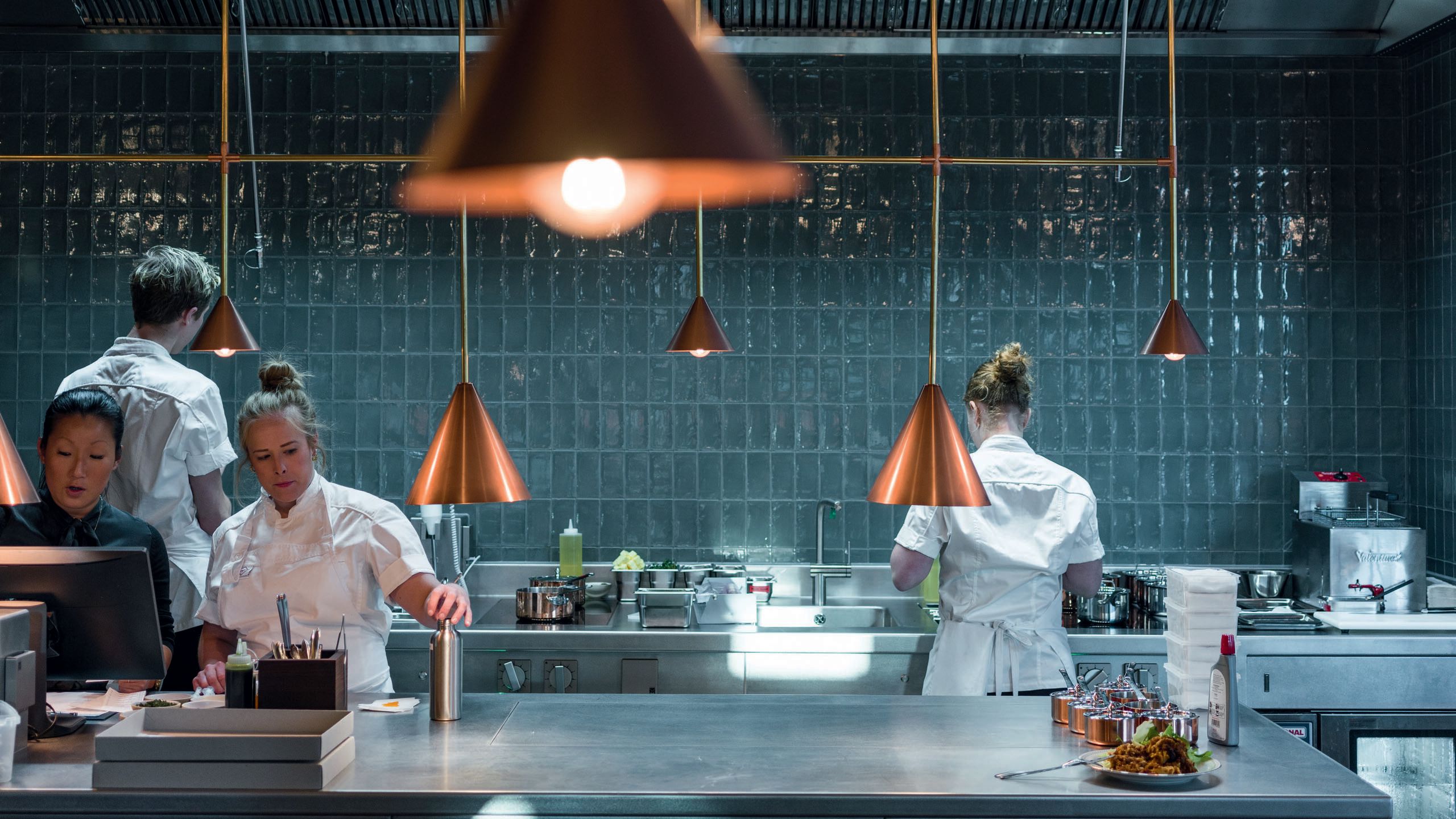 Inside the kitchen of the restairant - it is tiled in teal blue and has copper pendant lights. There are four people in the kitchen, three in chefs whites and a woman in a dark coloured top