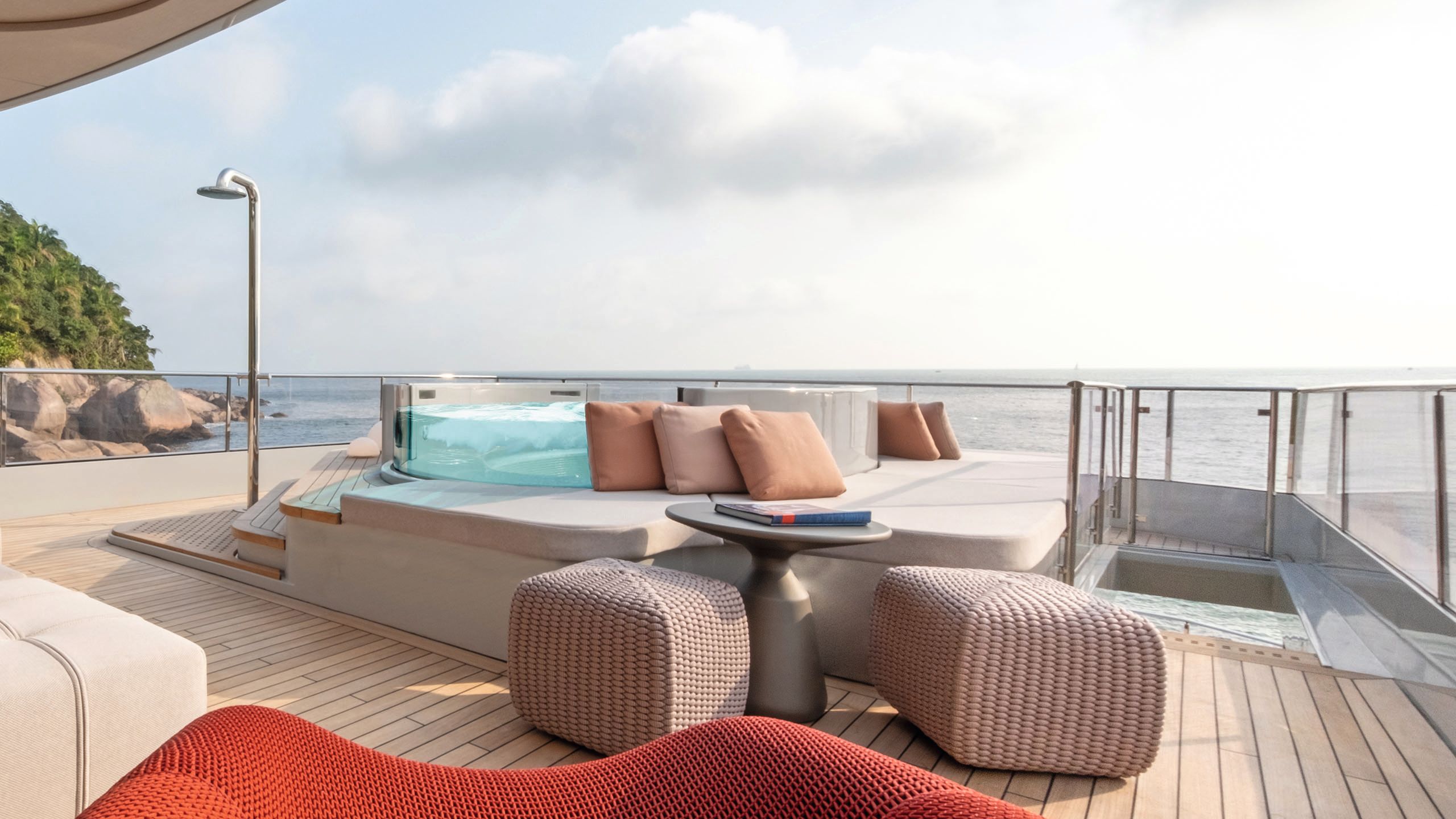 Side view of a platform with the glass-sided Jacuzzi in the centre. There is a sun pad surrounding it and soft furnishings in red and pink
