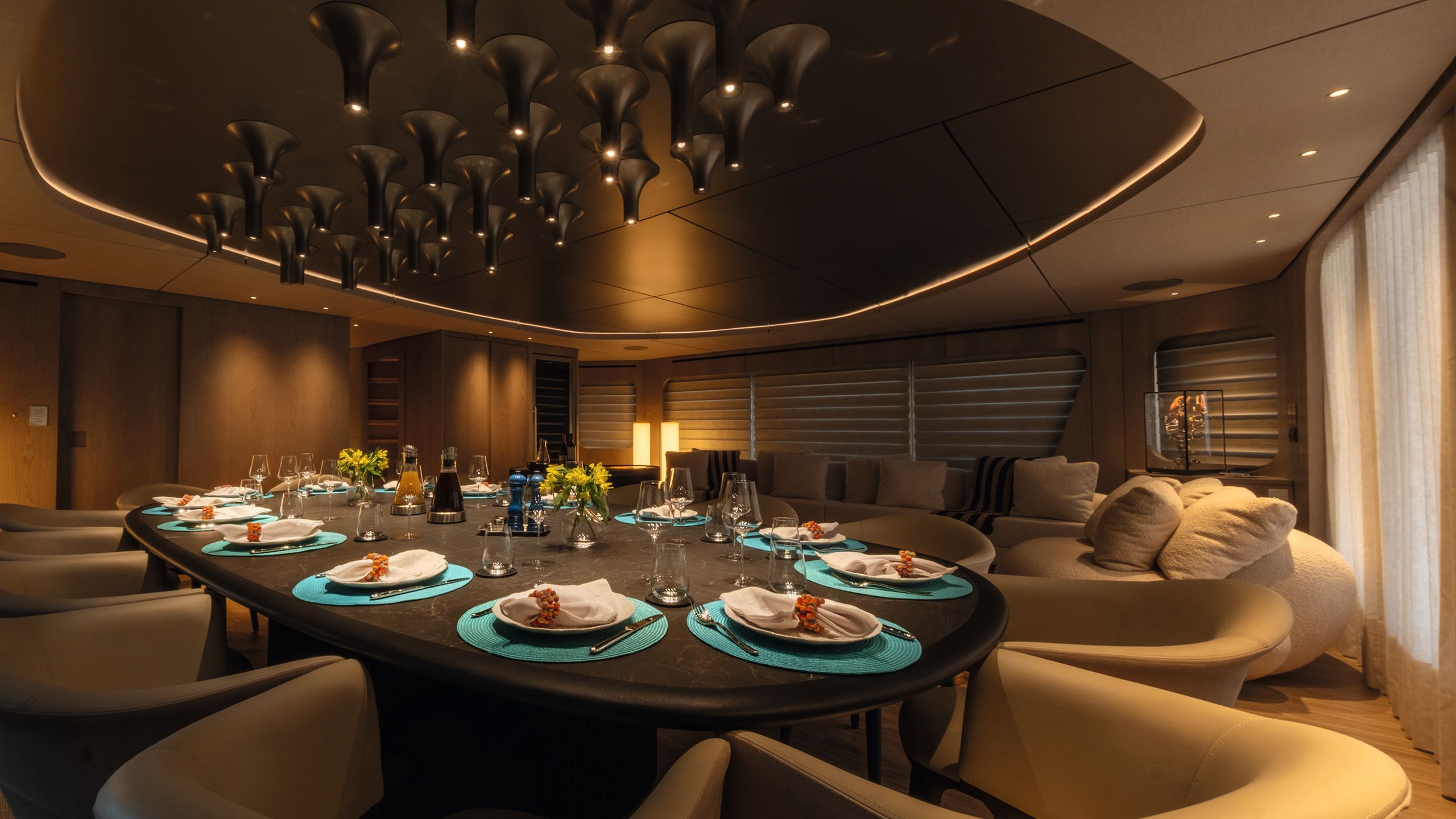 Alternative view of the dining table in the evening. The room is darkly lit and the table is set with turquoise place mats, white plates and cutlery. Overhead is a large lighting centrepiece made from abstract tubes