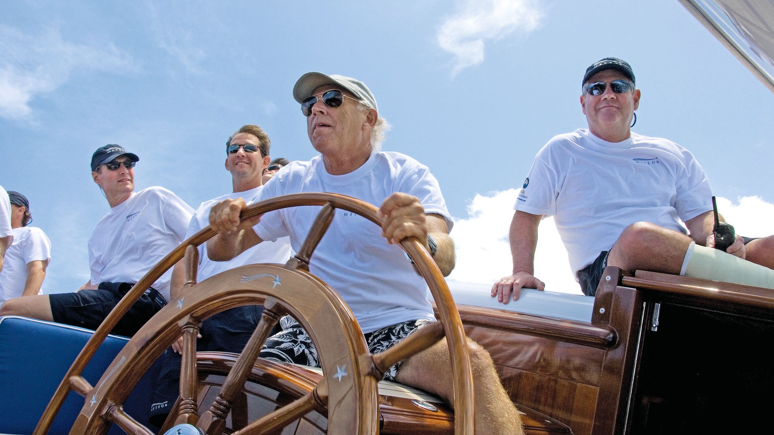 Jimmy Buffet is steering a boat. He is wearing a white t-shirt, beige cap, shorts and dark sunglasses. There are several other men sitting around him.