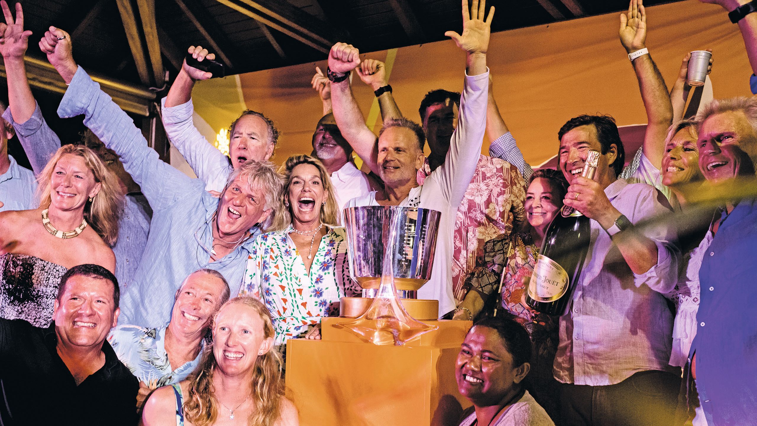 Group shot of crew members in front of a large silver trophy. They are all smiling with their arms raised in the air and one man is holding a large bottle of bubbly