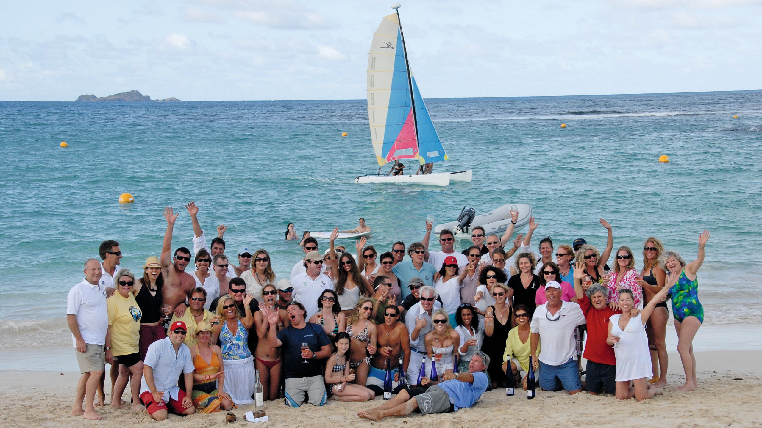 Group shot of about 60 people in beachwear and summer clothes on the beach. They're laughing and waving their hands in the air and looking at the camera.