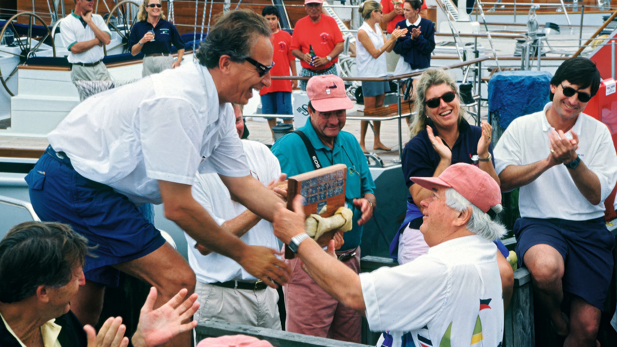 A man in a peach cap and white t-shirt hands an award to a man in a white shirt and blue shorts. They are both tanned.