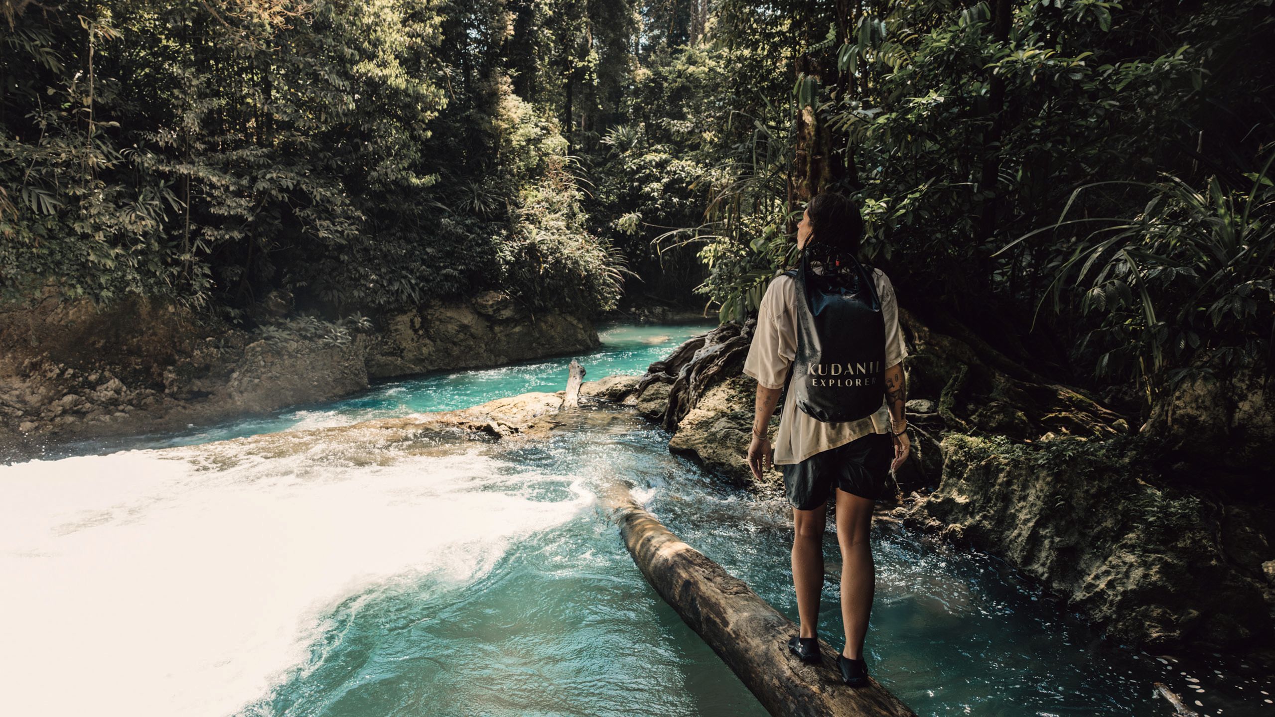 A woman in a t-shirt and shorts walking along a log crossing a stream in a forest