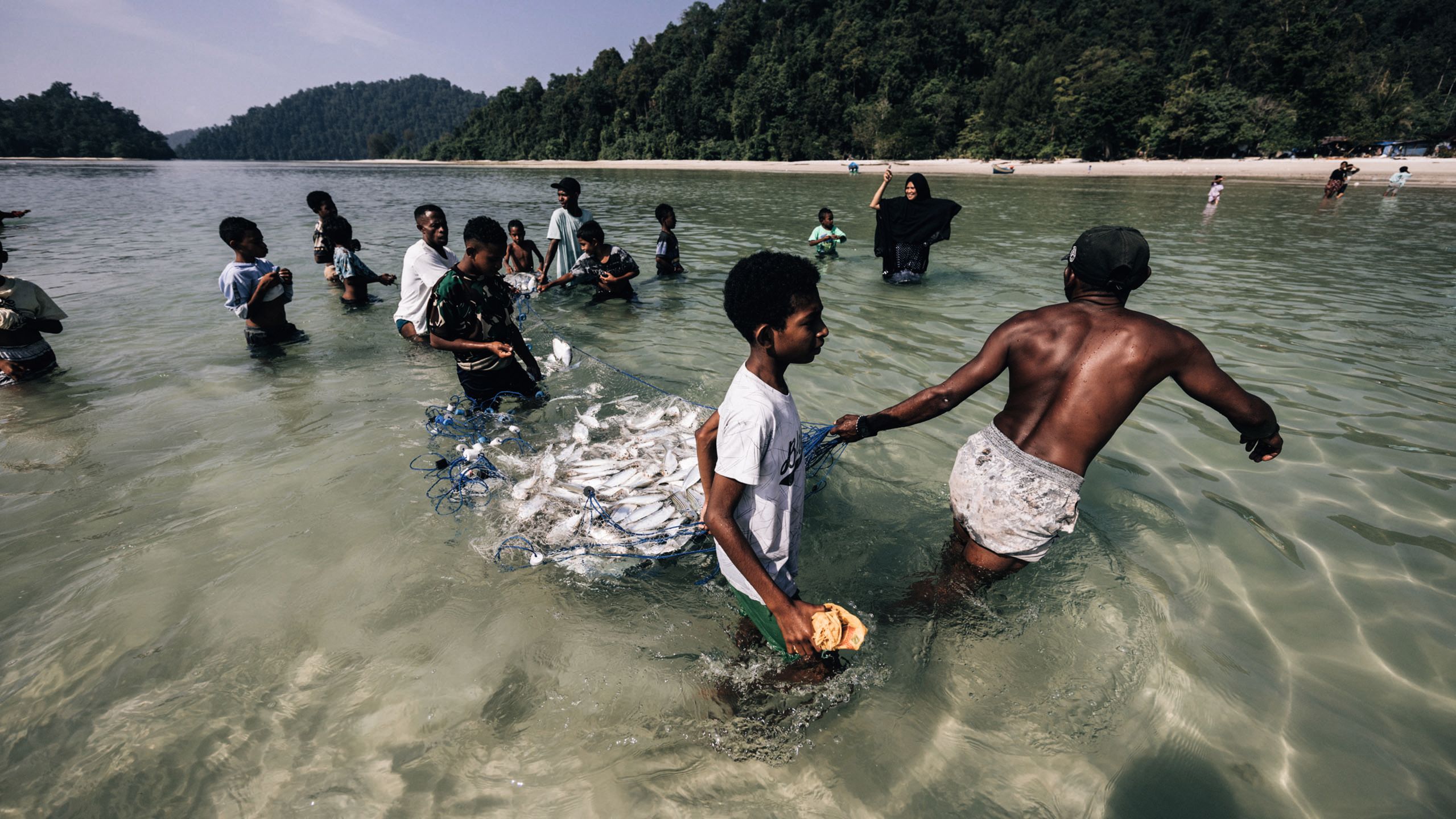 A group of about 12 men, both young and old, and one woman, standing in the shallows of the ocean hauling a huge net of fish in