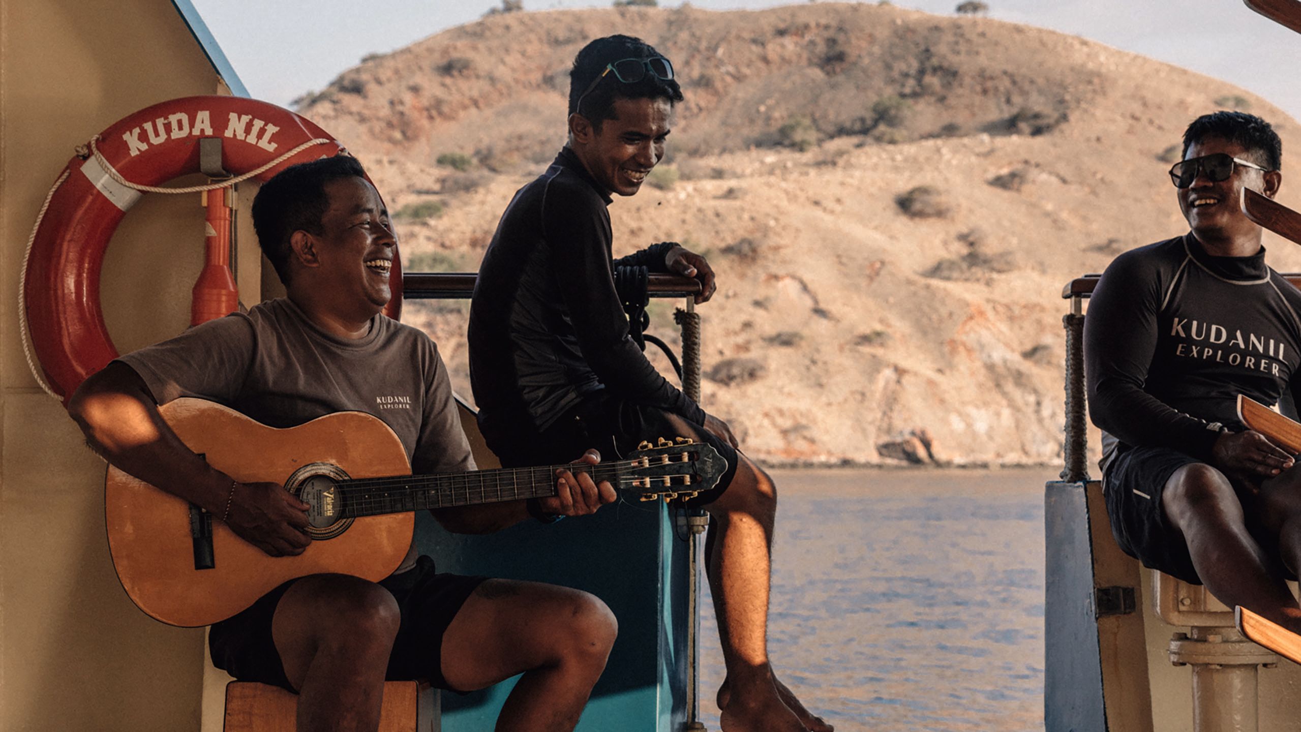 A man laughs while playing an acoustic guitar. Two other men are sat near him laughing as well