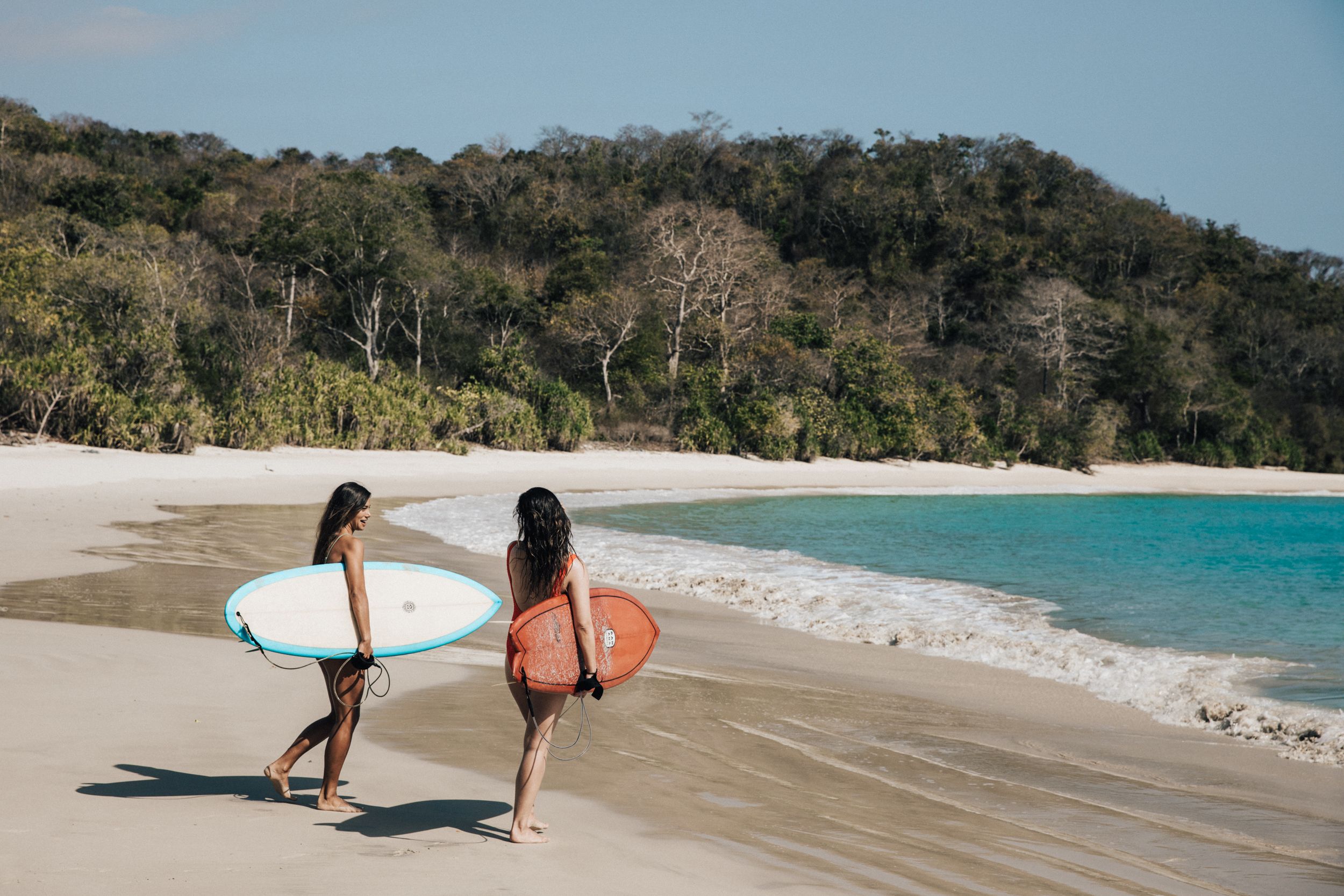 Two surfers with their back to the camera with boards under their arms, walking towards the shore at sunset