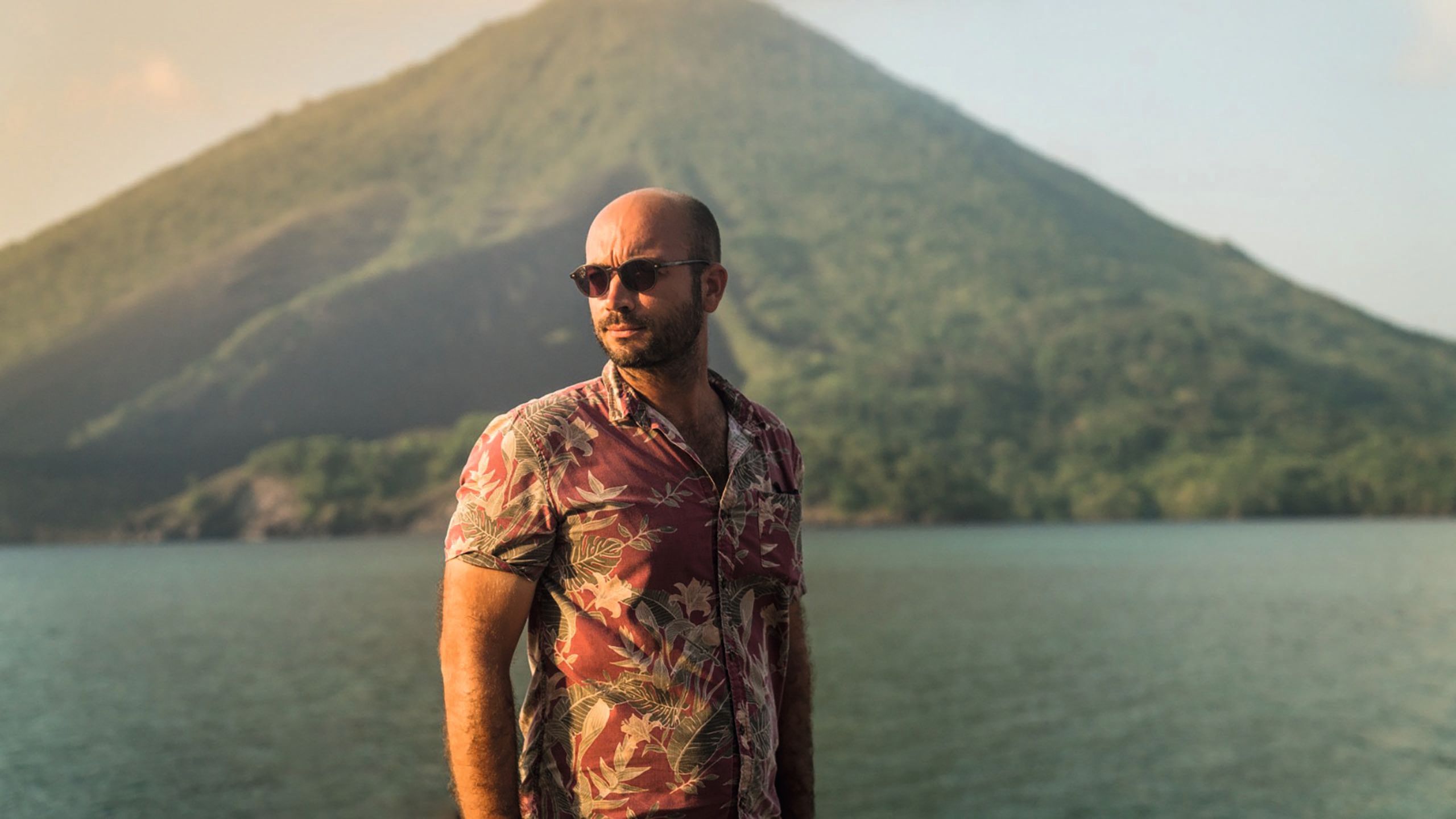 Thibaud Epstein shot from the waist up. He's looking off to the left and wearing a tight Haiwain shirt and sunglasses. A mountain and the ocean is in the background