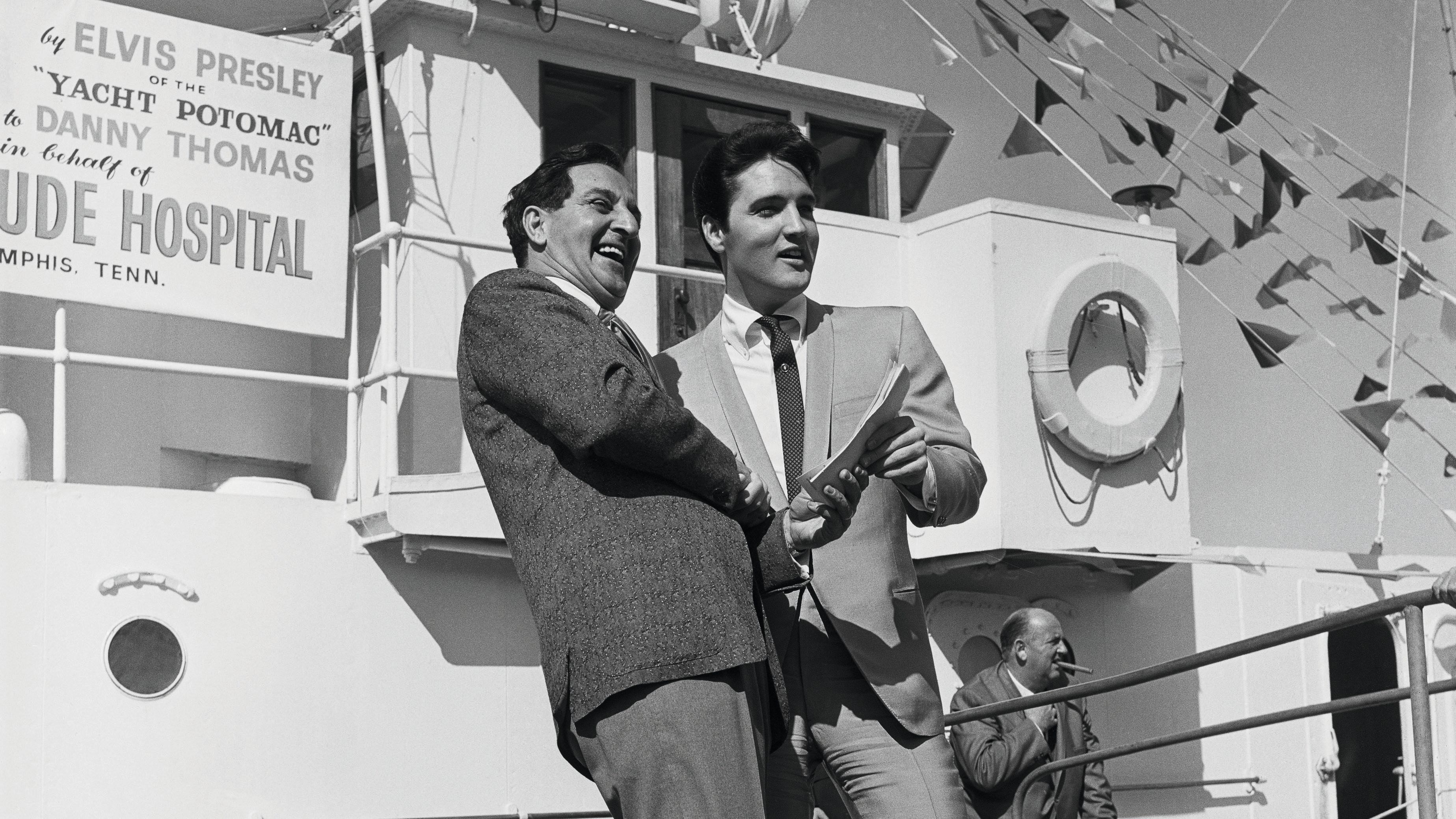 Presley is in a light grey suit and handing the papers to a man in a darker grey suit. There's bunting on the boat and a sign that says Potomac and Presley's name behind them