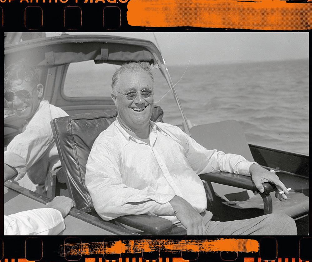 Black and white photo of FDR, sitting in a chair on the boat. He's wearing a white shirt, sunglasses, smiling at the camera and holding a cigarette