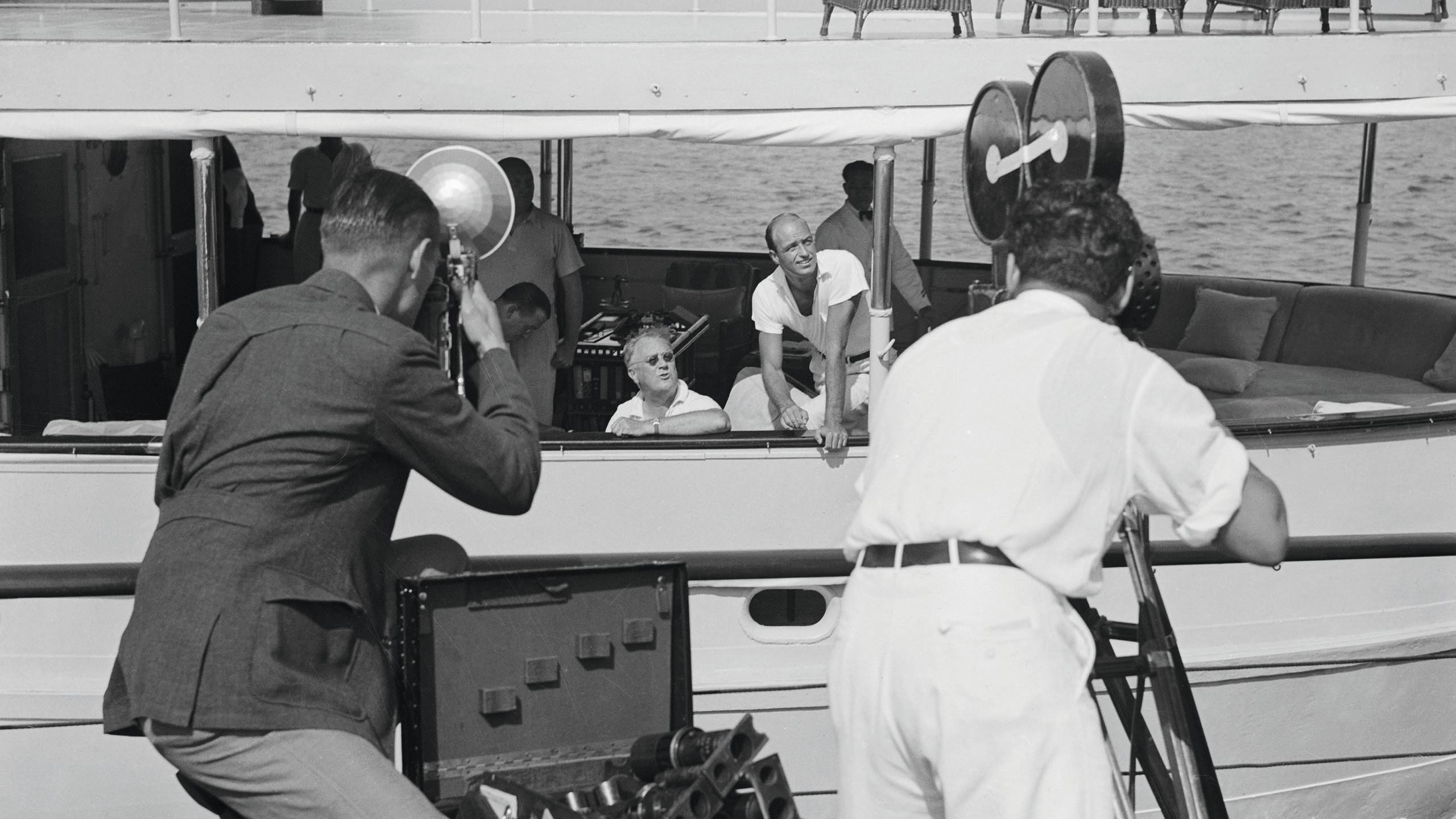 Two photographers are on the dock - one if filming FDR on the boat and another is holding a camera