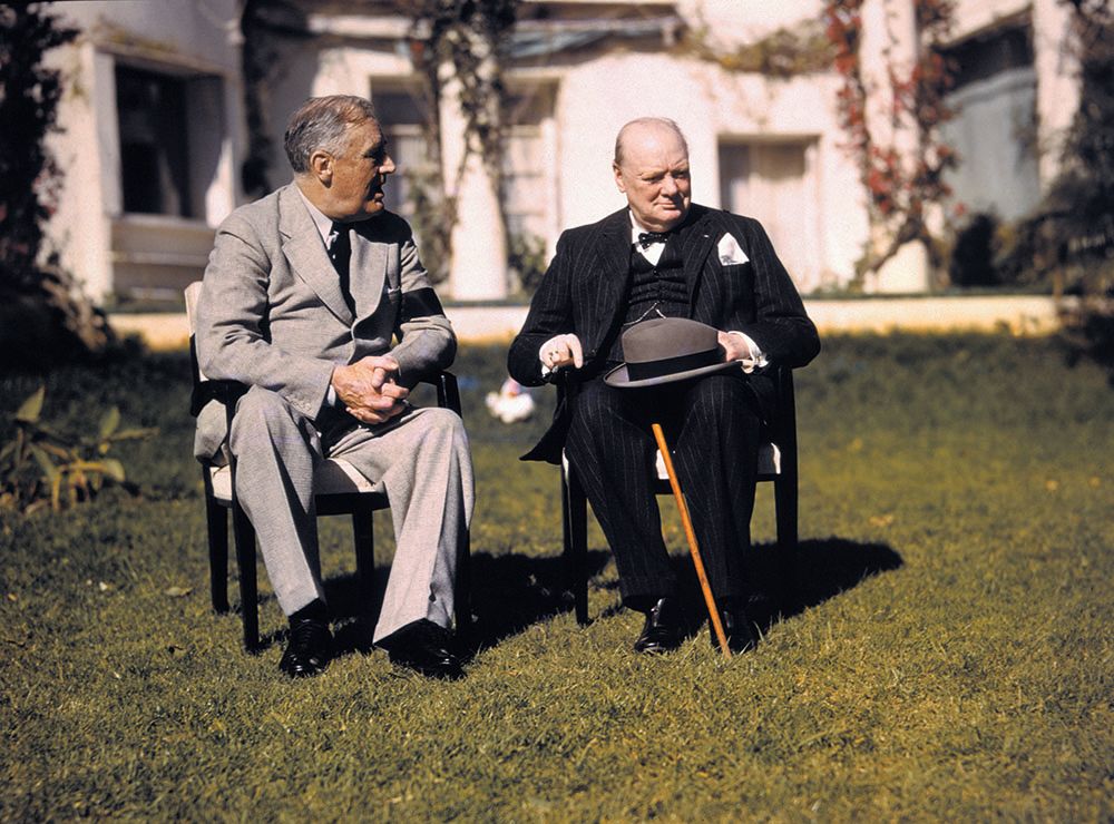 Roosevelt and Churchill are sitting in chairs, the former is wearing a grey suit and the latter is wearing a black suit with his hat and cane in his lap
